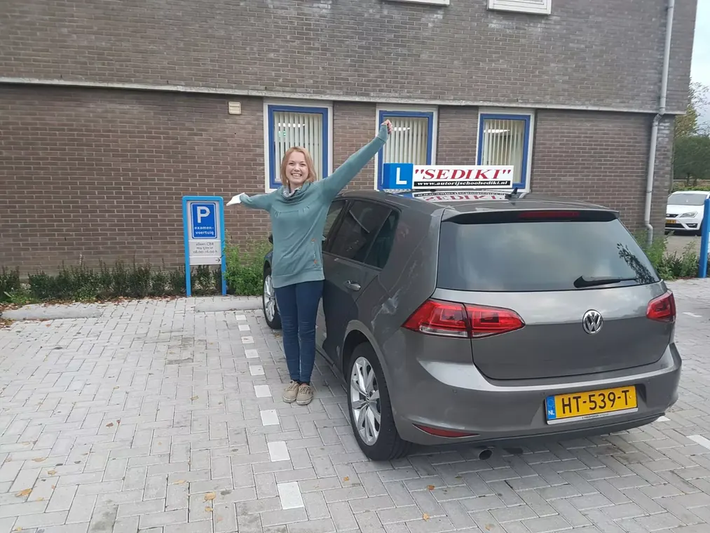 Geslaagde leerling van Autorijschool Sediki juicht naast de grijze VW Golf lesauto in Gouda